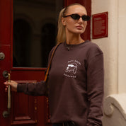 Woman wearing a brown sweatshirt with a graphic design, standing in front of a red door.